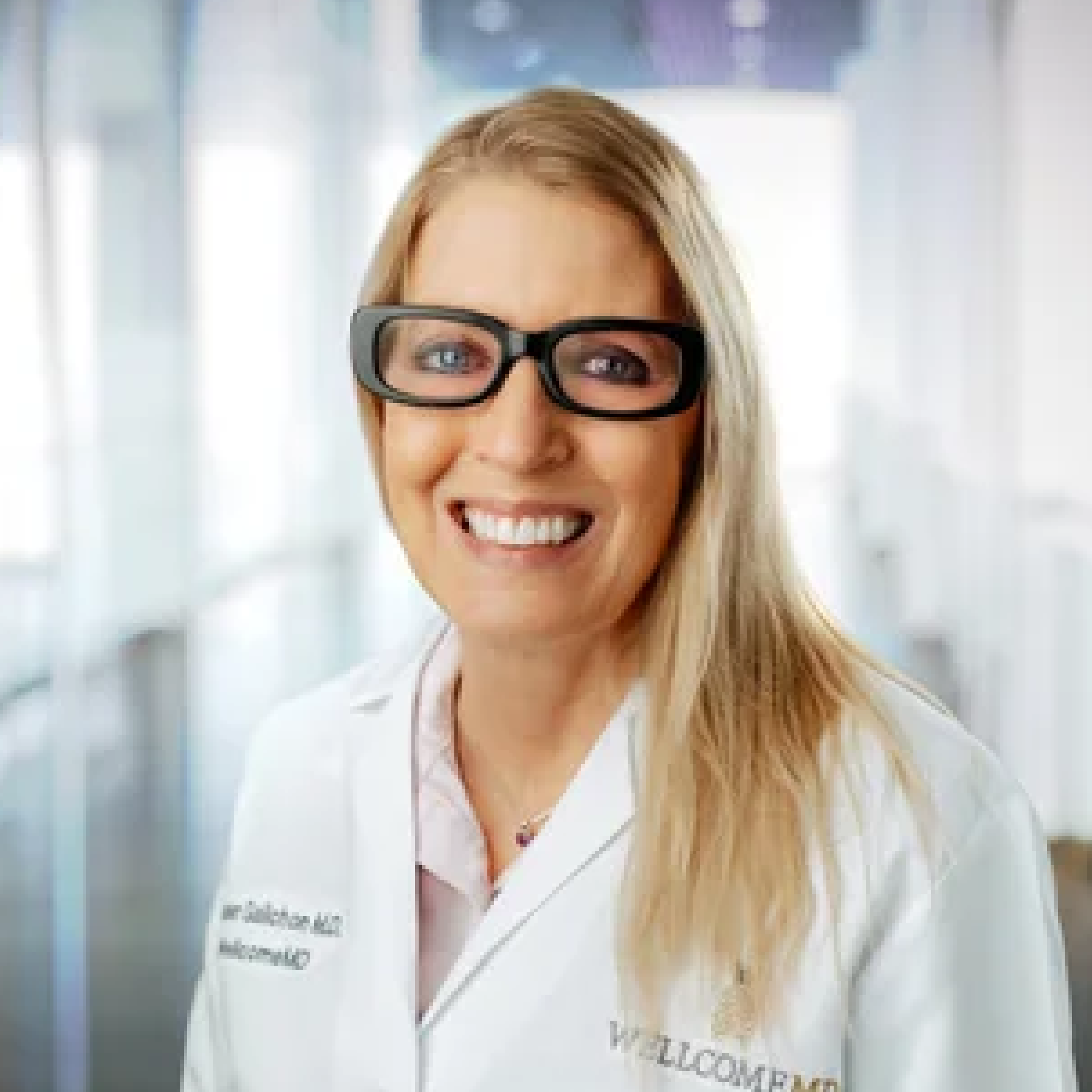Smiling woman with long blonde hair and black-framed glasses in a white lab coat.