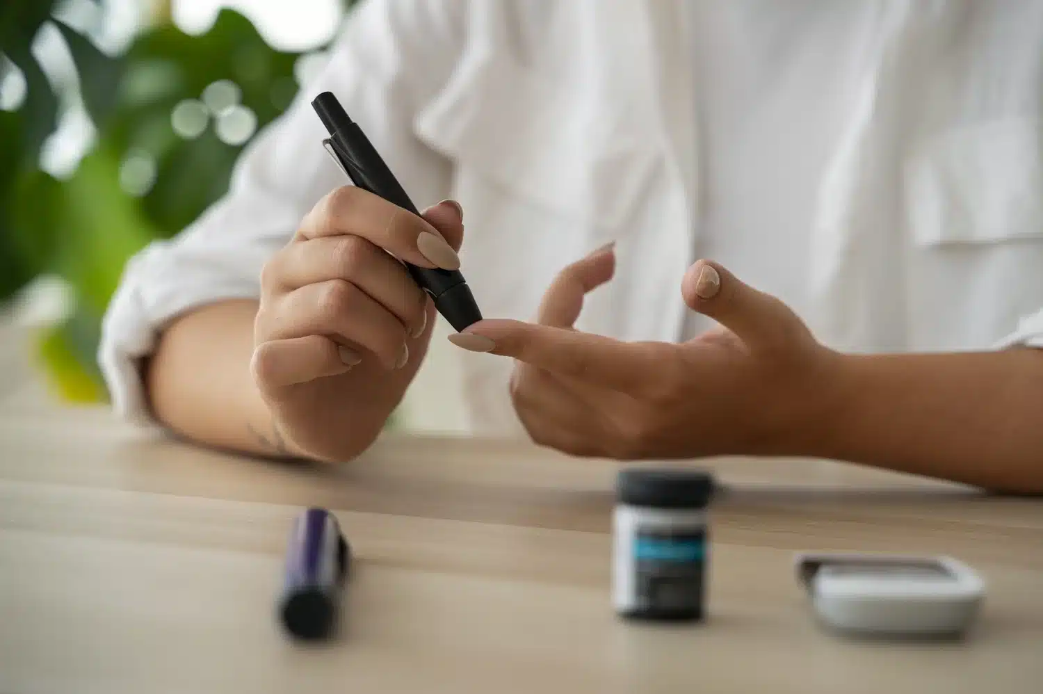 front-view-diabetic-woman-checking-her-glucose-level - WellcomeMD Person using a black lancing device to prick their fingertip for a blood glucose test, with testing supplies on the desk nearby.