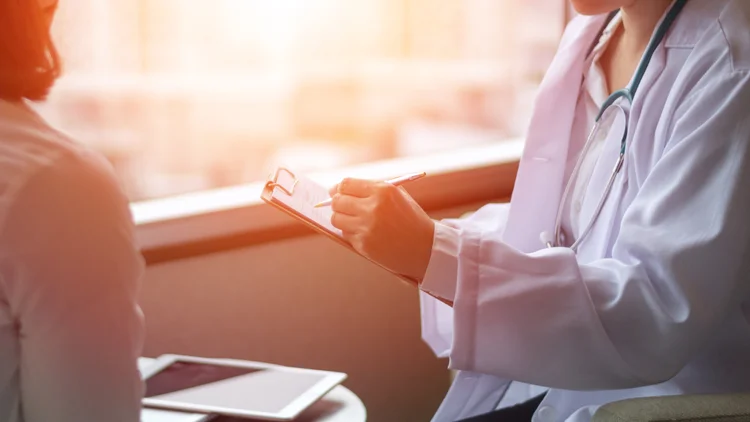 Doctor in a white coat with a stethoscope writing on a clipboard during a patient consultation.