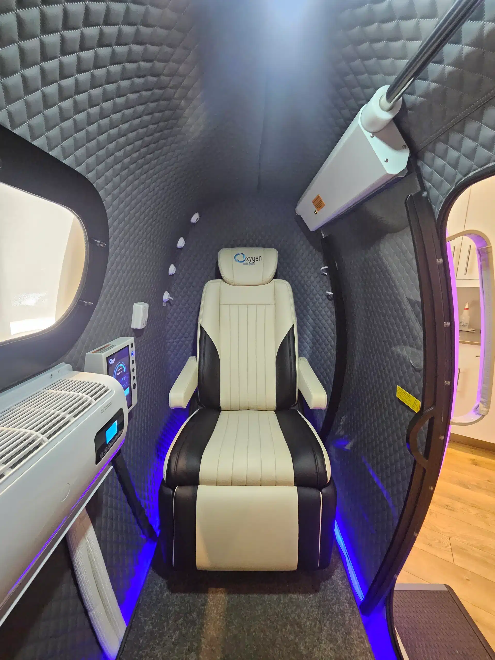HBOT Inside front view - WellcomeMD Interior of a compact medical capsule with quilted grey walls and a beige black seat labeled Oxygen, blue LED floor lighting, window on left, and equipment mounted along the wall.