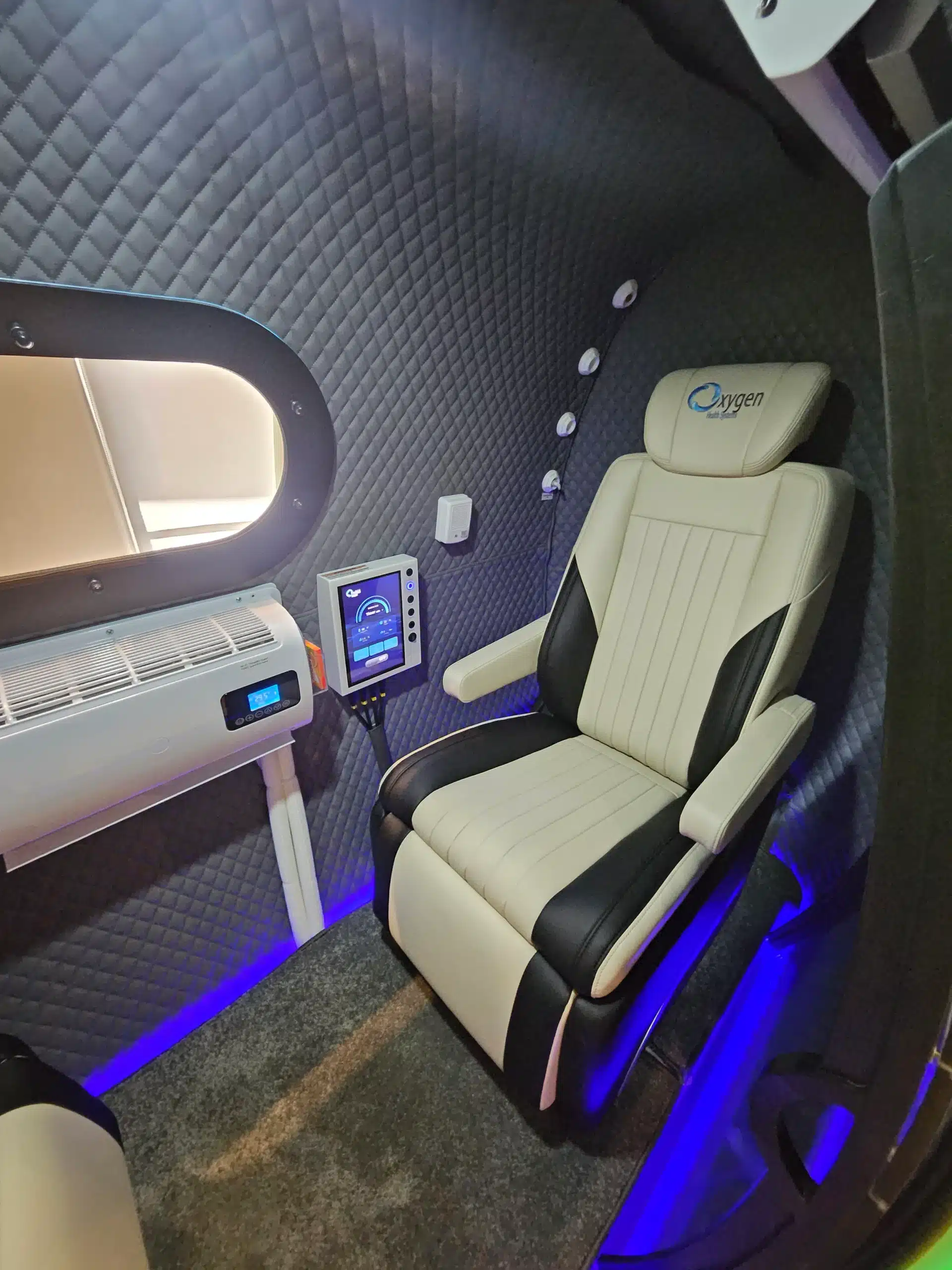Lower Page HBOT - WellcomeMD Luxurious pod interior with quilted dark walls, cream leather captain chair, medical/controls panel, and blue ambient lighting near a window.