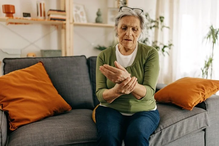 Elderly woman on a gray sofa in a bright living room, examining or massaging her wrist with a focused expression.