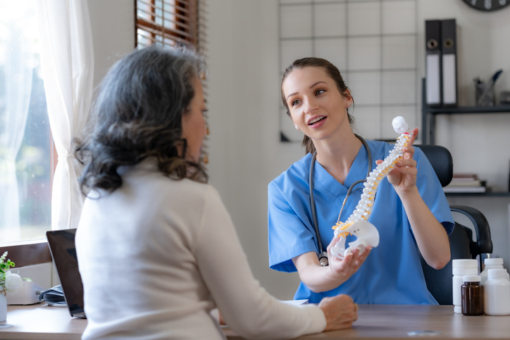 Spinal hygiene presentation Healthcare professional in blue scrubs shows spine model to patient in a clinic setting, explaining spinal anatomy at a desk with bottles nearby